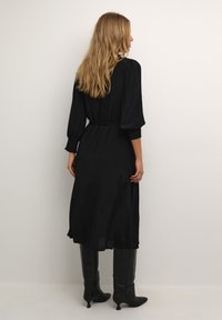 Zwarte jurk met lange mouwen, een cinched taille, een gladde textuur en een vloeiende rok. Gecombineerd met zwarte laarzen tot de knie.