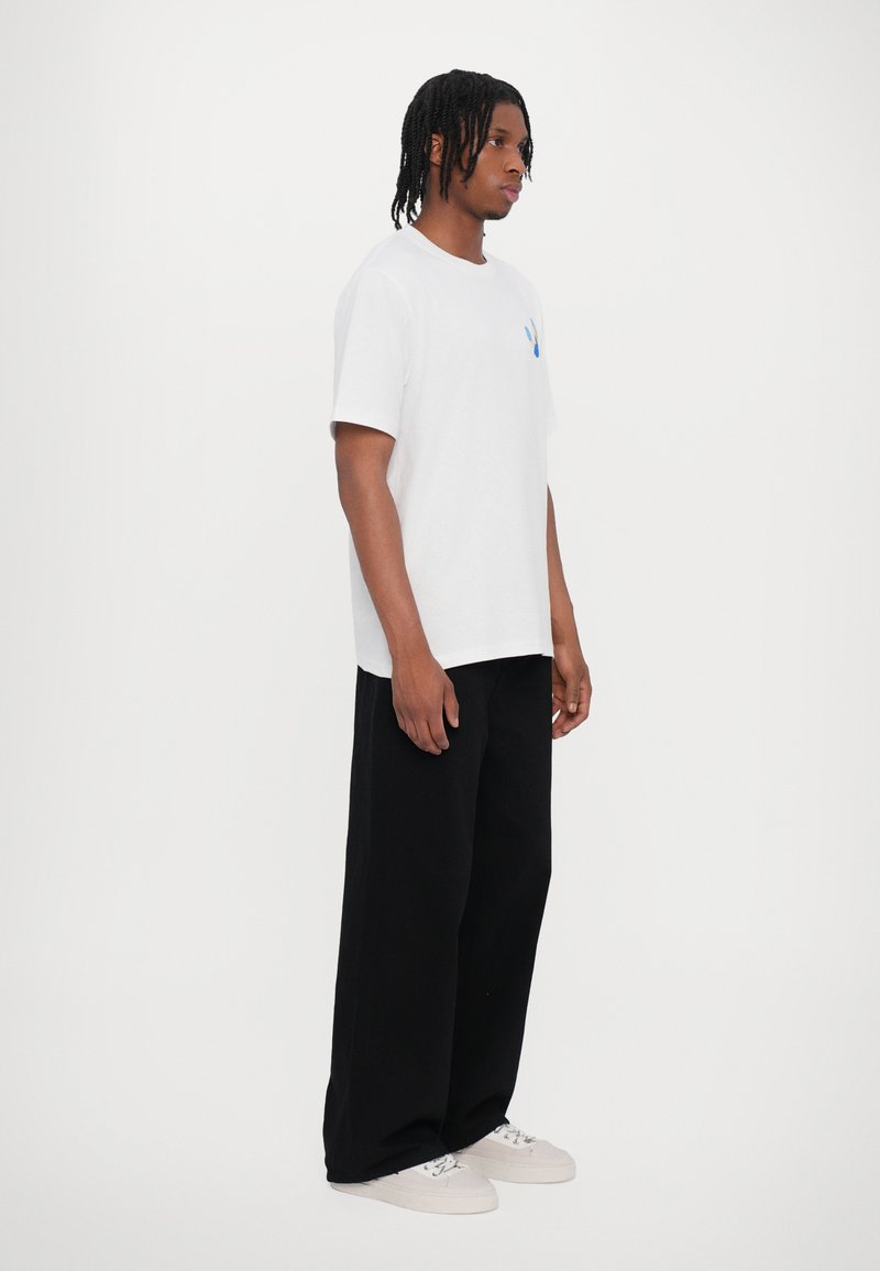 White cotton t-shirt with a blue accent on the chest, paired with loose-fitting black trousers and white sneakers.
