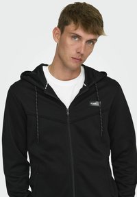 Veste à capuche zippée noire en tissu doux avec un cordon de serrage, présentant un patch logo blanc contrastant sur la poitrine gauche et une doublure texturée.