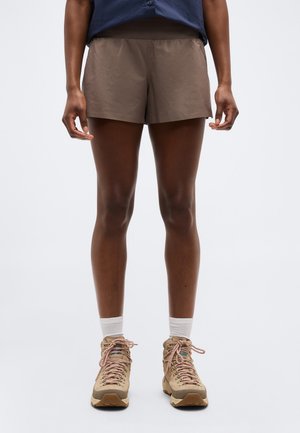 Persoon draagt bruine korte broek, beige wandelschoenen met roze veters, witte sokken, staande tegen een effen lichte achtergrond.