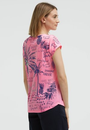 Frau mit hellbraunen Haaren trägt ein pinkes Kurzarmshirt mit navyblauen tropischen Prints und Text, kombiniert mit dunkler Hose, seitlich gewandt.