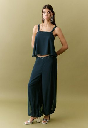 Jeune femme portant un haut sans manches noir et un pantalon à jambes larges, avec des talons argentés et des boucles d'oreilles pendantes, debout devant un fond beige uni.