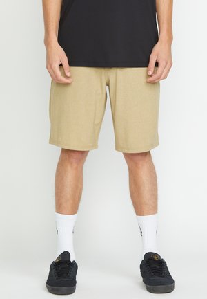 Homme portant un short beige, des chaussettes blanches montantes, des baskets noires et un t-shirt noir, debout devant un fond clair uni.