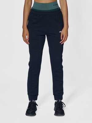 Femme portant un pantalon de jogging noir, des chaussures de sport noires et une ceinture taille haute turquoise, debout devant un fond uni.