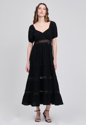 Frau trägt ein schwarzes Midikleid mit Puffärmeln, Spitzeneinsätzen und einem Rüschensaum, kombiniert mit schwarzen, rückenfreien Sandalen mit hohen Absätzen, vor einem grauen Hintergrund.