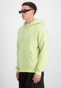 Felpa verde chiaro con cappuccio e coulisse, dotata di tasca frontale, realizzata in tessuto morbido; presenta un piccolo logo bianco sul petto e orlo a coste.