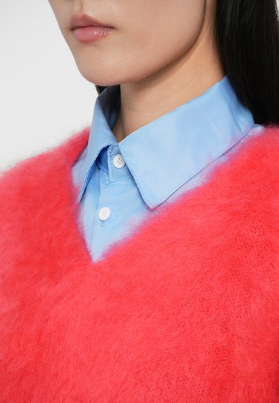 Maglione rosa fluffoso con scollo a V indossato su una camicia azzurro chiaro con bottoni, caratterizzata da una superficie liscia e colletto a punta.