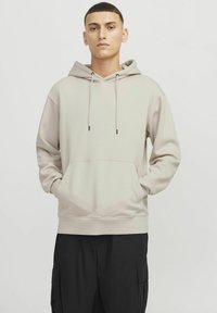 Cream hoodie with drawstrings and front pocket. Features a relaxed fit and ribbed hem. Worn with loose black cargo pants.
