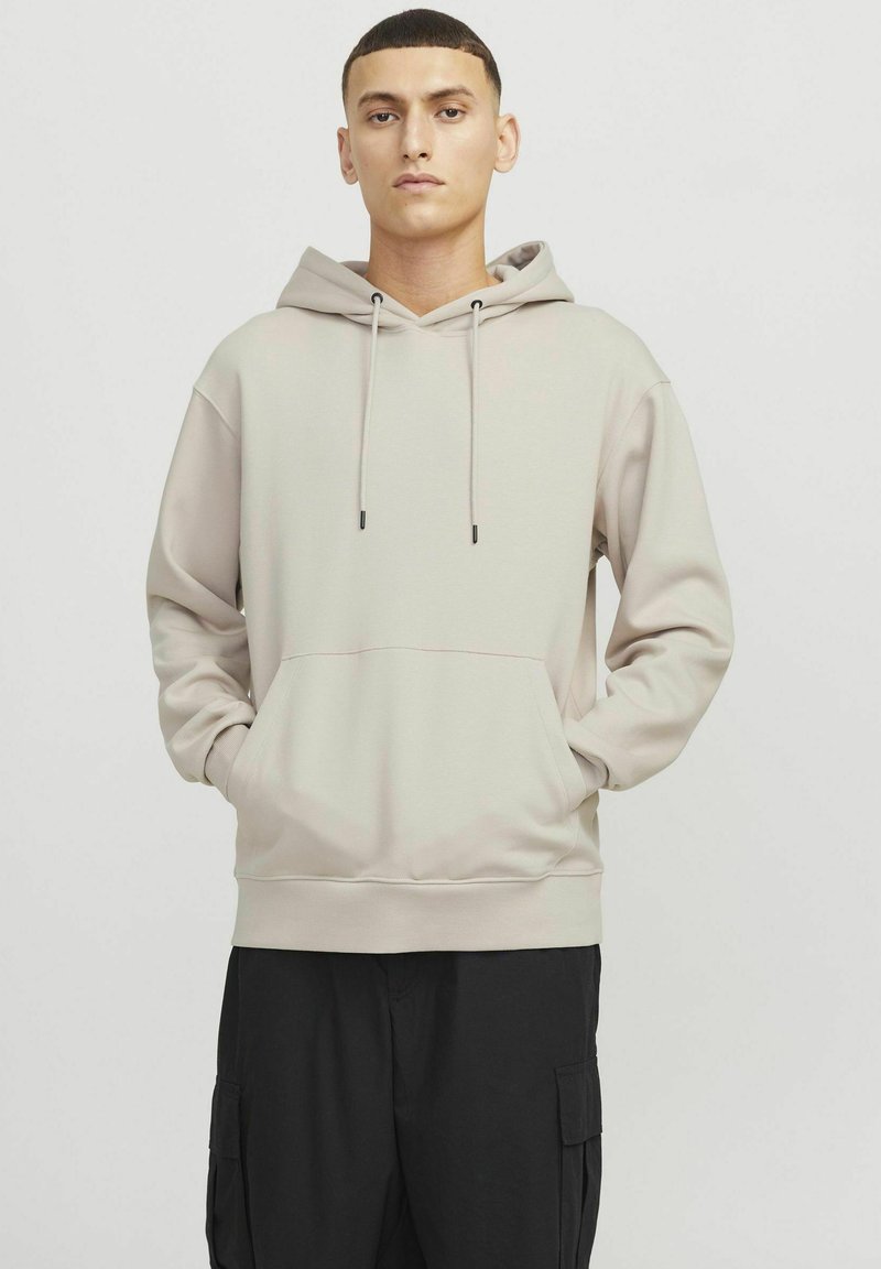 Cream hoodie with drawstrings and front pocket. Features a relaxed fit and ribbed hem. Worn with loose black cargo pants.