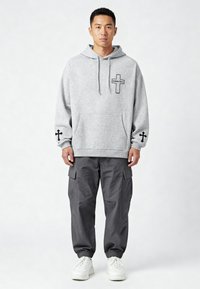 Felpa grigia con cappuccio e design a croce neri, tasca a marsupio e dettagli neri sulle maniche. Abbinata a pantaloni cargo grigi scuri e scarpe da ginnastica bianche.
