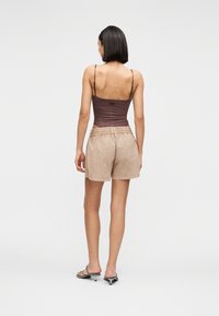 Γυναίκα με μαύρα κοντά μαλλιά φοράει καφέ αμάνικο τοπ, μπεζ σορτς και μαύρες σανδάλες με τακούνι, στέκεται με την πλάτη προς τα εμπρός σε απλό φόντο.