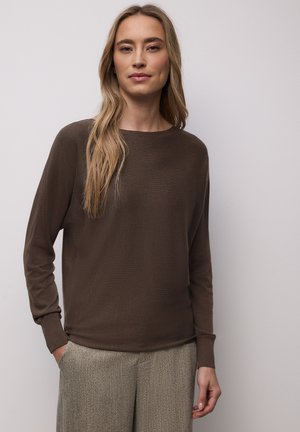 Žena s dlouhými blond vlasy, oblečená v hnědém tričku s dlouhými rukávy a šedých kalhotách s texturou, stojící před jednobarevnou bílou zdí.