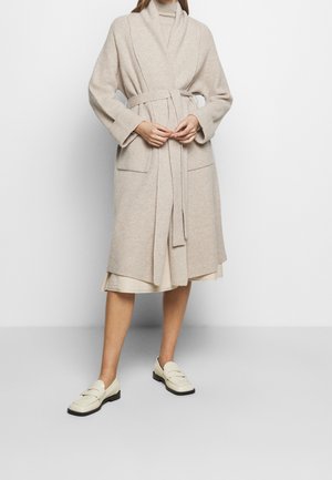 Nainen, joka pitää beigeä polvipituista neulotua neuletakkia vyöllä kermaisen mekon ja kermaisten loafereiden päällä, seisoo valkoista taustaa vasten.