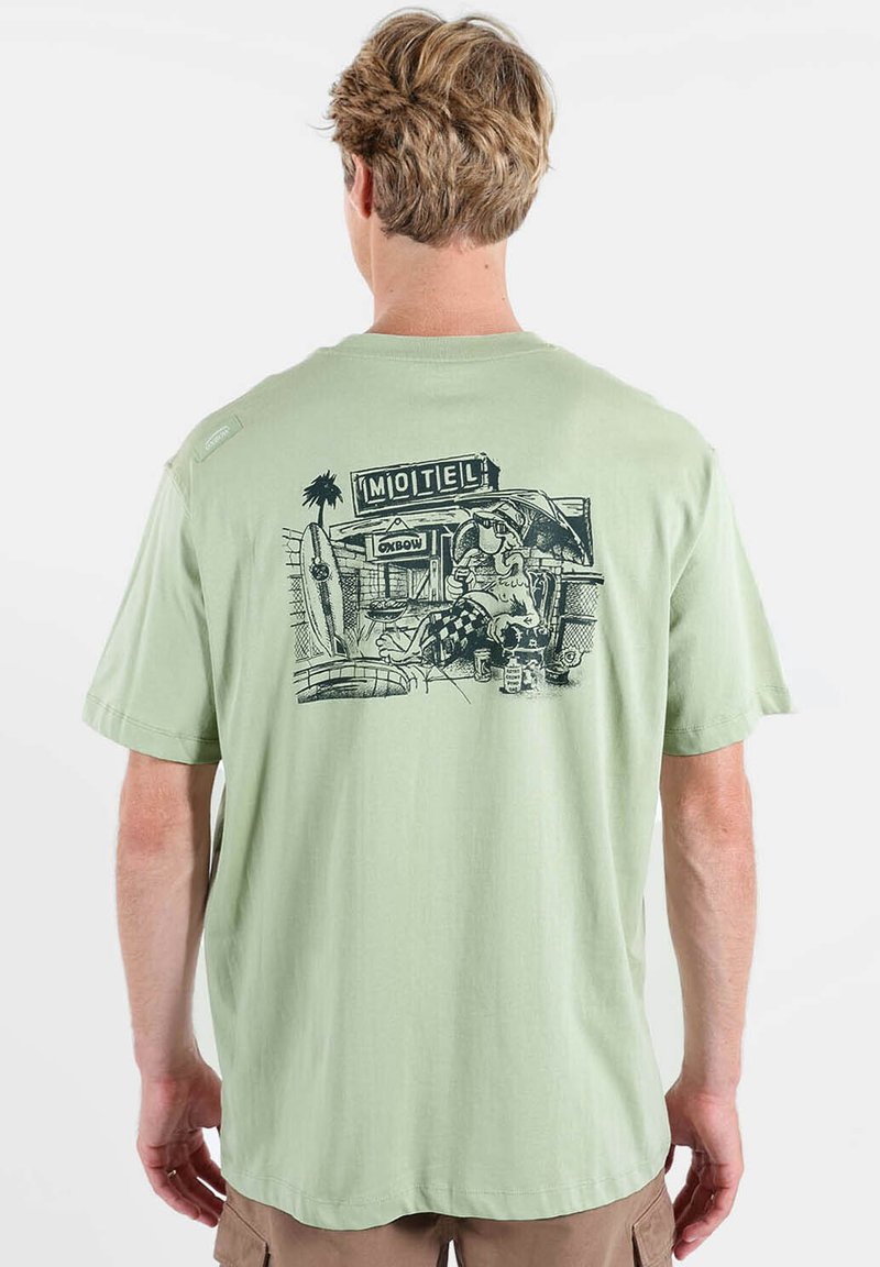 T-shirt en coton vert clair avec une impression graphique noire au dos représentant une scène de motel avec des planches de surf et un personnage détendu.