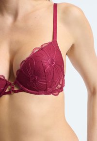 Reggiseno in pizzo borgogna con ricami floreali, caratterizzato da pannelli trasparenti, bordi ondulati e spalline regolabili con chiusura a gancio.
