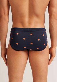 Slip de bain marine avec un motif de petits logos de super-héros rouges et jaunes. Le tissu semble lisse avec un design ajusté.