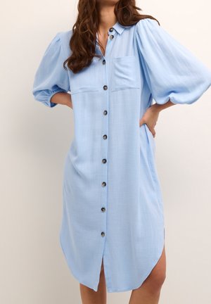 Vestido camisero azul claro con mangas abullonadas y bolsillos delanteros en el pecho, usado por una mujer con las manos en las caderas.