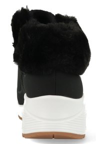 Zapatilla negra con un suave cuello de pelo sintético negro, entresuela blanca y suela de goma color caramelo. Superficie lisa con detalles de costura visibles.