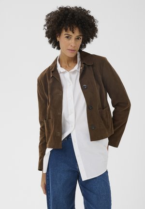 Frau mit lockigem Haar, die eine braune, geknöpfte Jacke über einem weißen Hemd und blauen Jeans trägt, vor einem schlichten hellen Hintergrund steht.