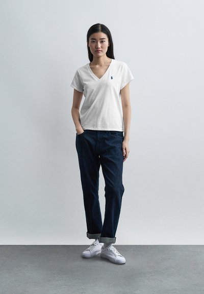 Νεαρή γυναίκα που φοράει λευκό T-shirt με V λαιμόκοψη, σκουρόχρωμα τζιν καμπάνα, και λευκά αθλητικά παπούτσια, στέκεται μπροστά σε απλό γκρι φόντο.