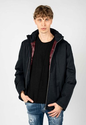Mørkeblå jakke med lynlås foran, hætte og burgunderfor. Har et afslappet design og struktureret stof. Bæres over en sort T-shirt.