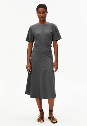 Woman wearing a fitted, short-sleeve, dark gray midi dress and black strappy flat sandals, standing against a plain white background.