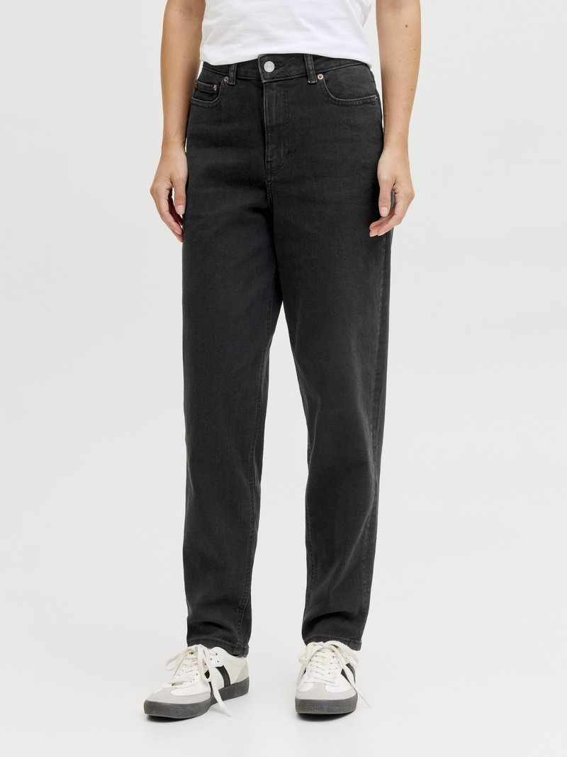 Person wearing high-waist black jeans and white sneakers with black stripe, standing with hands relaxed at sides on a plain background.