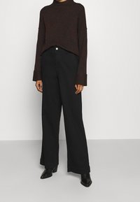 Tummanruskea neule, jossa on korkea kaulus ja pidennetyt hihat, yhdistettynä korkeavyötäröisiin mustiin leveälahkeisiin housuihin ja mustiin korkokenkiin.