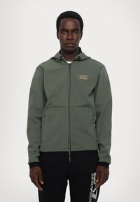 Giacca con cappuccio e zip di colore verde oliva, realizzata in tessuto liscio, con una tasca frontale e un piccolo logo dorato sul petto, abbinata a pantaloni neri.