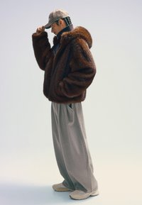 Brun fuskpäls oversize-jacka med huva, grå plisserade byxor och beige sneakers, kompletterat med en rutigt keps.