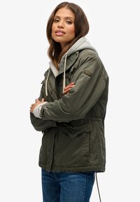 Olivengrøn jakke med hætte, der har lynlås foran, snøre i taljen og ribkant ved ærmerne, lagdelt over en grå hættetrøje. Denimjeans synlige.