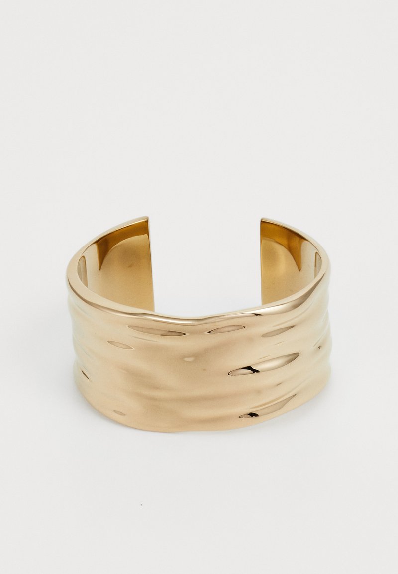 Kuldne käevõru lainelise kujundusega, sile pind ja avatud otsad. Peegeldav pind näitab õrnu kõverusi ja delikaate valguse peegeldusi.