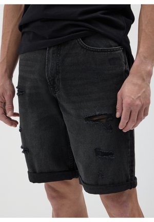 Mann trägt schwarze, abgenutzte Denim-Shorts und ein schwarzes Hemd, steht mit entspannt an den Seiten hängenden Händen.