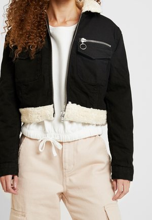 Femme portant une veste courte noire avec un bord en polaire blanche, un sweat-shirt court blanc et un pantalon beige clair à cordon.