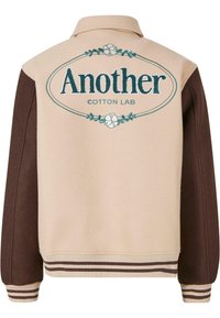 Jachetă bej și măcărlău cu un design cu guler. Prezintă un text brodat îndrăzneț "Another Cotton Lab" și detalii florale. Manșete și tiv cu dantelă.