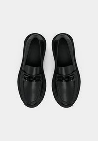 Sapatos slip-on de couro preto com biqueira arredondada, apresentando um detalhe de corrente preta grossa e solas texturizadas. Design minimalista, acabamento suave.