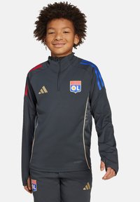 Veste d'entraînement zippée noire avec des rayures bleues et rouges sur les épaules, accents dorés et logo de l'Olympique Lyonnais. Présente une coupe athlétique et des manches longues.