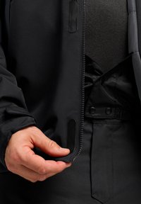 Veste noire imperméable dotée d'une fermeture éclair sur le devant et d'une fermeture à pression. Tissu texturé avec des poignets élastiques, mettant en avant une main ajustant l'ourlet.