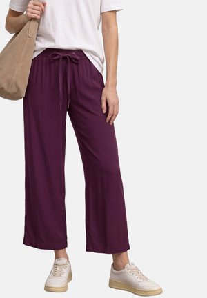 Osoba nosząca luźne fioletowe spodnie ze sznurkiem, białe trampki, białą koszulkę i nosząca beżową zamszową torbę na ramię.