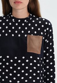 Camicia bianca e nera a pois con scollatura rotonda e una tasca quadrata marrone sul lato sinistro. Tessuto morbido, maniche lunghe.