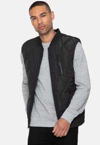 Gilet nero trapuntato con chiusura a zip, colletto a costine e tasca laterale con zip, indossato sopra un felpa grigia. Tessuto a superficie liscia.