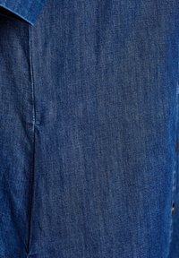 Jeans tyg i djupblå färg med synlig textur. Har sömdetaljer och knappackenteringar längs ena sidan.