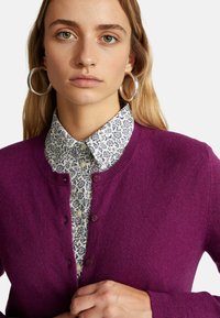 Fialový pletený kardigan s kulatým výstřihem, zapínáním na knoflíky a žebrovanými manžetami, vrstvený přes bílé květinové košilové tričko.