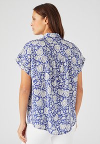 Camisa de manga corta de tejido azul con patrón floral blanco, que cuenta con un cuello y un pliegue en la espalda para mayor libertad de movimiento.