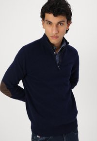 Pulover u tamnoplavoj boji s polu-zip ovratnikom, teksturiranim materijalom i smešnim laktovima, uparen s košuljom s uzorkom ispod.
