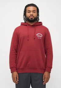 Hombre con rastas que lleva una sudadera con capucha roja con bolsillo frontal y pequeño texto blanco en el lado izquierdo del pecho, de pie contra un fondo blanco.