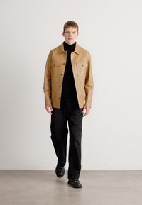 Καφέ δερμάτινο μπουφάν με δύο θηκάκια στο στήθος, κουμπί κλεισίματος και λεία υφή, σε συνδυασμό με μαύρο πουλόβερ και μαύρο παντελόνι.