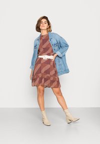 Casaco de ganga sobre um vestido estampado em vermelho escuro com um cinto branco. Botas de tornozelo bege. O vestido apresenta uma bainha com babados e padrões geométricos variados.