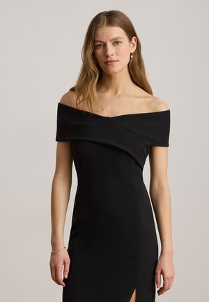 Kvinde med langt, lysebrunt hår iført en tætsiddende sort off-shoulder kjole med en frontslids, stående mod en ensfarvet lys baggrund.