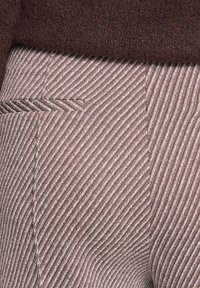 Gros plan sur un pantalon en laine rayé diagonal marron et crème avec une poche passepoilée et un bas en maille d'un pull marron foncé.